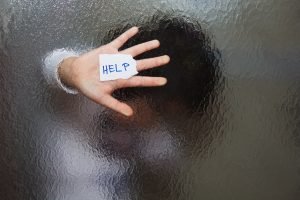 Woman standing behind blurred glass holding up a piece of paper that says 'help' symbolizing domestic violence at a home office. 